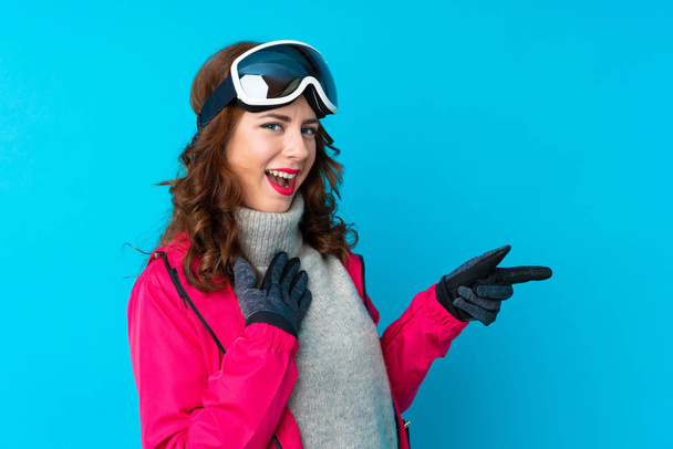 脇に指を指す隔離された青い壁の上にスノーボードメガネを持つスキーヤー女性 - 写真・画像