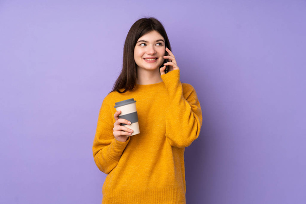 Young Ukrainian teenager girl over isolated purple background holding coffee to take away and a mobile - Фото, изображение
