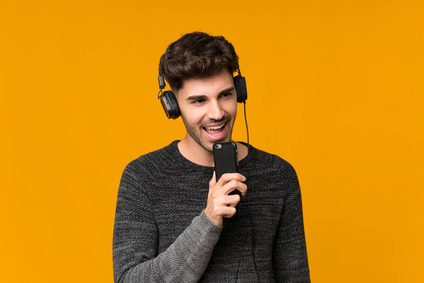 Young man over isolated background using the mobile with headphones and singing - Photo, Image