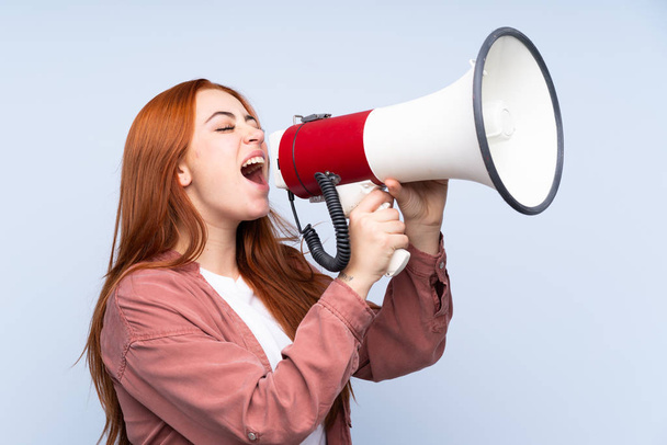 Рыжая девушка-подросток на изолированном синем фоне кричит через мегафон
 - Фото, изображение