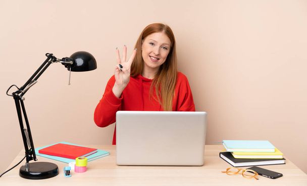 Young student woman in a workplace with a laptop happy and counting three with fingers - Photo, Image