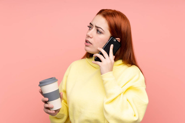Redhead teenager girl over isolated pink background holding coffee to take away and a mobile - Фото, изображение