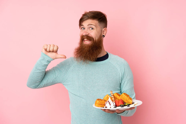 Redhead man with long beard holding waffles over isolated pink background proud and self-satisfied - Valokuva, kuva