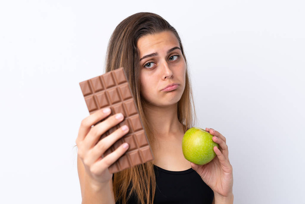 Teini-ikäinen tyttö yli eristetty valkoinen tausta ottaen suklaa tabletti toisessa kädessä ja omena toisessa
 - Valokuva, kuva