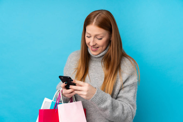 Genç kızıl saçlı kadın, mavi arka planda, elinde alışveriş çantaları ve cep telefonuyla bir arkadaşına mesaj yazıyor. - Fotoğraf, Görsel