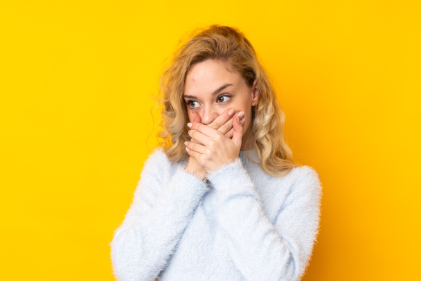 Young blonde woman isolated on yellow background covering mouth and looking to the side - Foto, imagen