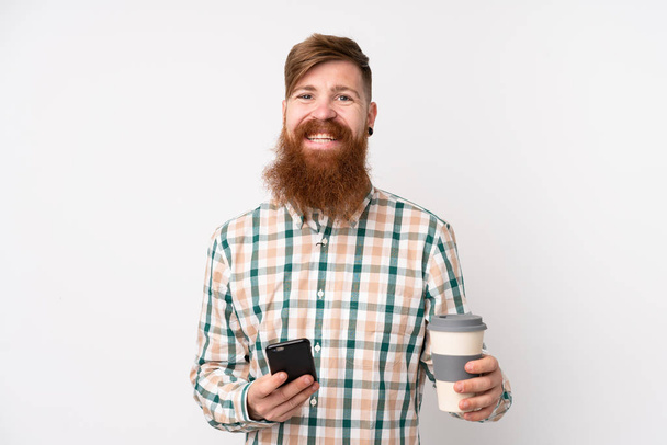 Redhead man with long beard over isolated white background holding coffee to take away and a mobile - Photo, Image
