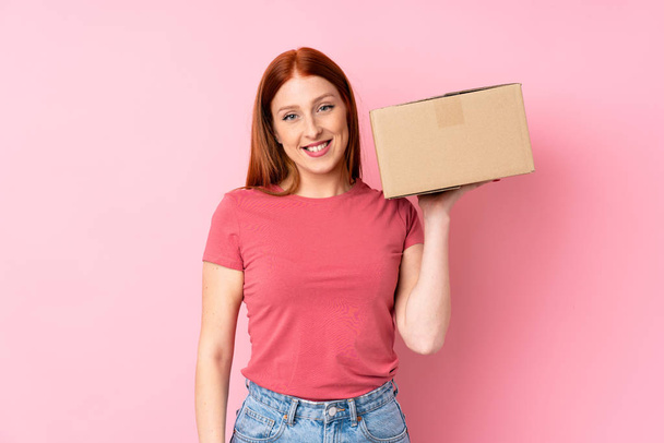 Молода руда жінка над ізольованим рожевим тлом тримає коробку, щоб перенести її на інший сайт
 - Фото, зображення