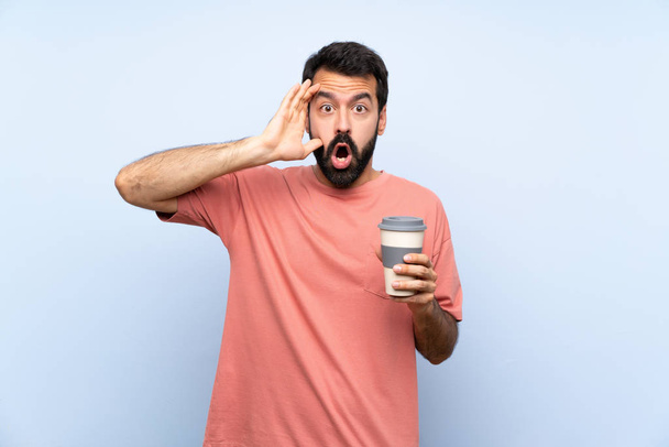 Jeune homme barbe tenant un café à emporter sur fond bleu isolé avec expression surprise
 - Photo, image