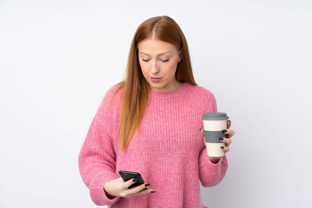 Joven pelirroja con suéter rosa sobre fondo blanco aislado sosteniendo café para llevar y un móvil - Foto, imagen