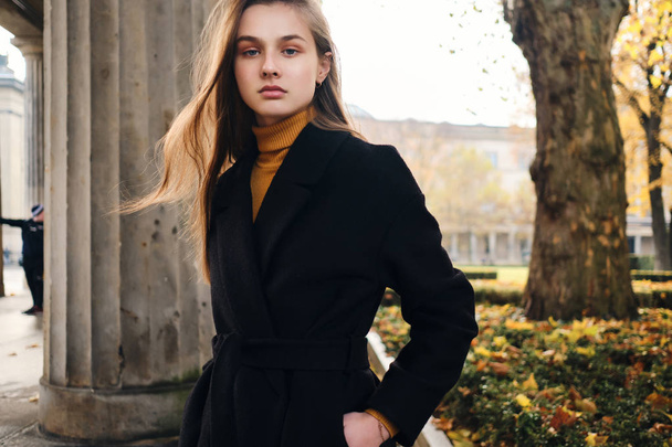Beautiful serious girl in coat confidently looking in camera in autumn city park - Foto, afbeelding