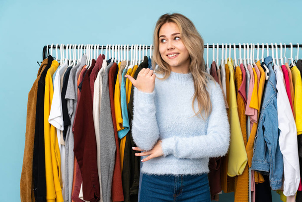 十代のロシアの女の子は、製品を提示する側を指している青の背景に孤立したいくつかの服を購入 - 写真・画像