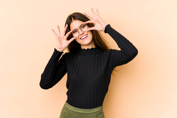 Young caucasian woman isolated on beige background showing okay sign over eyes - Foto, afbeelding