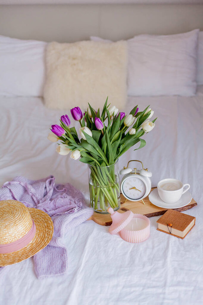 Een boeket tulpen in een glazen vaas, een witte wekker, een kaars, een briefje, een cappuccino mok op een houten plank staan op het bed. Vrouwenspullen. Ontbijt op bed. - Foto, afbeelding
