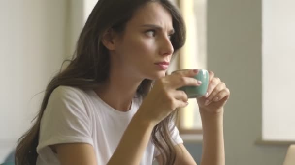 Concentrated serious young woman is drinking coffee while working with a laptop indoors - Video