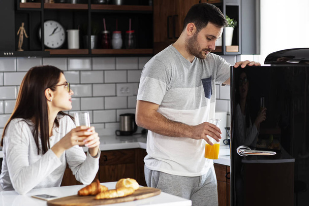 Couple in the kitchen eat breakfast with orange juice and pastry at table and he open refrigerator - Photo, image