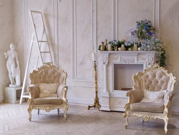 Δύο μπεζ vintage καρέκλες με χρυσή πέτρα στο εσωτερικό με φόντο ένα τζάκι διακοσμημένο με πεύκα στεφάνια και κεριά, στο βάθος είναι ένα άγαλμα και μια ξύλινη σκάλα - Φωτογραφία, εικόνα