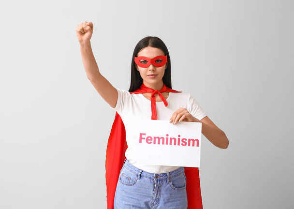 Süper kahraman kostümü giymiş genç bir kadın elinde gri arka planda feminizm metni tutan bir kağıt tutuyor. - Fotoğraf, Görsel