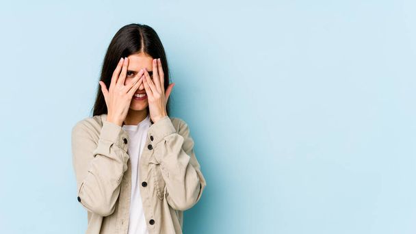 Νεαρή καυκάσια γυναίκα απομονωμένη σε μπλε φόντο ανοιγοκλείνει τα μάτια της στην κάμερα μέσα από δάχτυλα, ντροπιασμένη καλύπτοντας το πρόσωπο. - Φωτογραφία, εικόνα