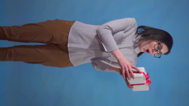 happy young business woman with a gift box in her hands stands on a blue background vertical video - Felvétel, videó