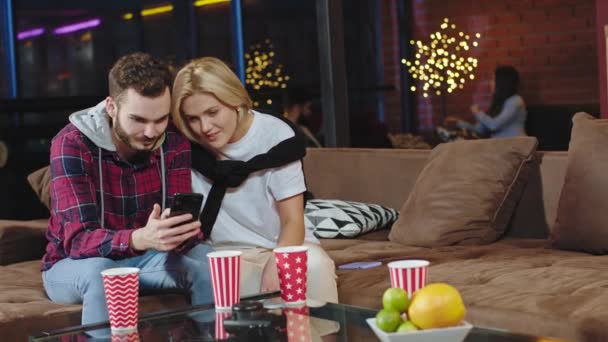 Blonde lady with short hair and charismatic guy smiling large they sitting on the sofa and enjoying the time together looking through the phone and chatting - Záběry, video