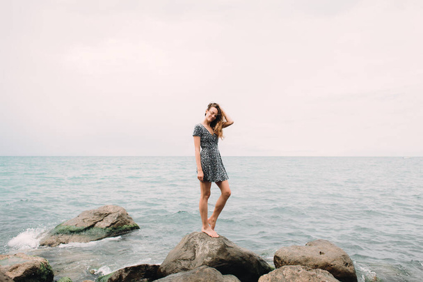egy fiatal lány a ruha áll a strandon nagy kövek, a szél fejleszti a haját - Fotó, kép