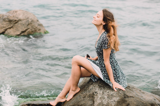 a young beautiful girl in a dress is sitting on large rocks near the sea, the wind is developing her hair - Fotoğraf, Görsel