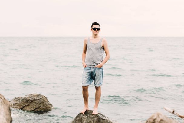 a young guy in a t-shirt and shorts stands on the beach on large rocks - Φωτογραφία, εικόνα