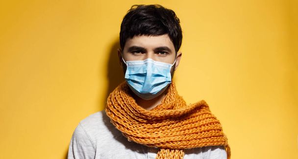 Portret van een jonge man met een blauw griepmasker, met oranje sjaal, geïsoleerd op gele achtergrond. - Foto, afbeelding