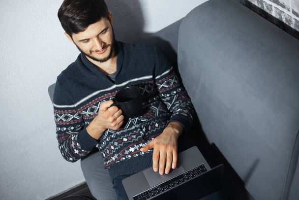 Портрет молодого уверенного в себе человека с черной кружкой в руке, сидящего дома на диване и использующего ноутбук
. - Фото, изображение