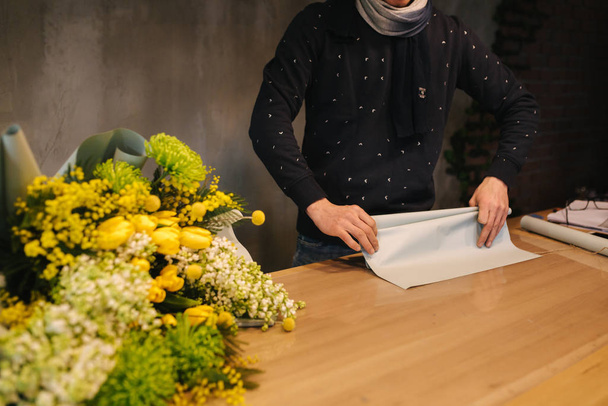 Mężczyzna zrobić wiosenny bukiet. Człowiek kwiaciarnia zawijając piękny bukiet wiosennych kwiatów w papier paczkowy na drewnianym stole. Piękny kwiat kompozycja żółtych kwiatów - Zdjęcie, obraz
