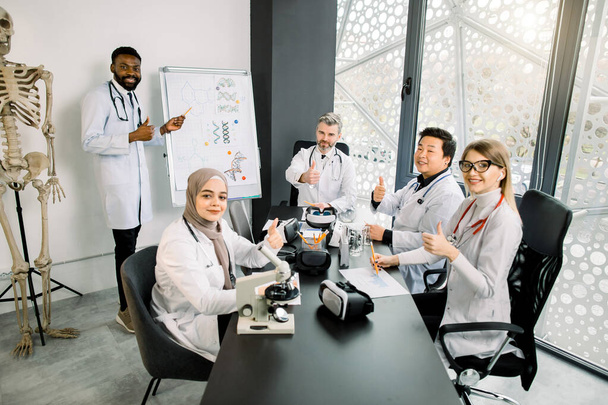group of multiethnic students, Caucasian and Muslim girls, African and Asian guys, and middle aged man professor teacher working at the laboratory, sitting at the table and showing thumbs up - Φωτογραφία, εικόνα