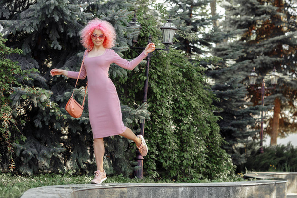 Dancing young woman with curly pink hair and sunglasses near the blossoming spring tree. - Φωτογραφία, εικόνα
