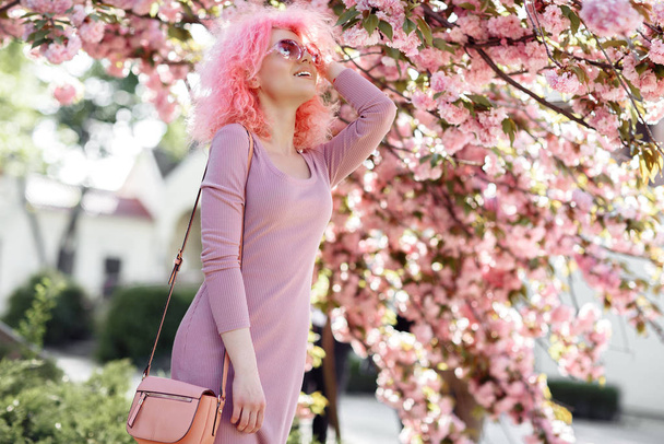 Positive young woman with curly pink hair and sunglasses near the blossoming spring tree. - Foto, Bild