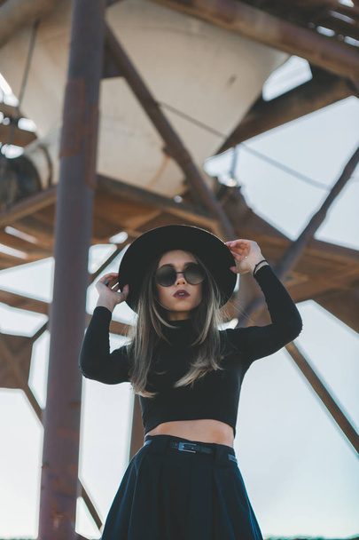 A long haired blonde woman, dressed all in black, wearing a black fedora type hat, black sun glasses and a knee high black dress, posing nonchalantly in front of a distressed industrial silo, in the m - Фото, зображення