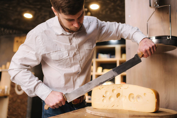 Young worker slice lactose-free cheese maasdam on wooden board in cheese shop - Photo, Image