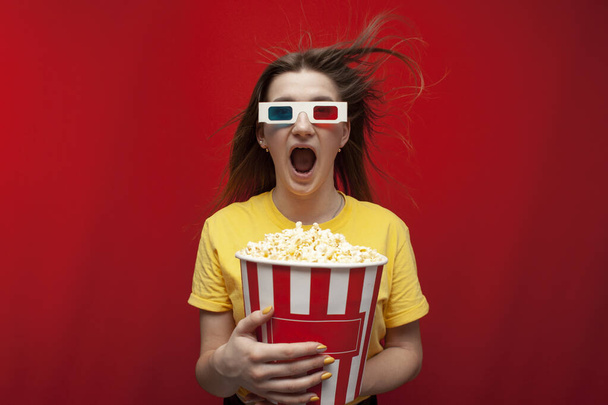 funny young girl student watching a movie in 3D glasses and screaming on a red background, she eats popcorn and is delighted - Photo, Image