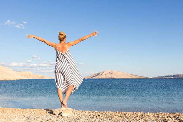 Boldog gondtalan nő felemelkedő karok, visel gyönyörű csíkos nyári ruha élvezi késő délután a fehér zavaros strand Pag sziget, Horvátország - Fotó, kép