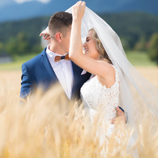 Sposo abbraccia teneramente sposa mentre il vento soffia il suo velo nel campo di grano da qualche parte nella campagna slovena
. - Foto, immagini