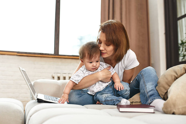 Žena s dítětem u laptopu, sedící na pohovce. Práce doma, na volné noze, práce během mateřské dovolené pro vzdálený přístup. - Fotografie, Obrázek
