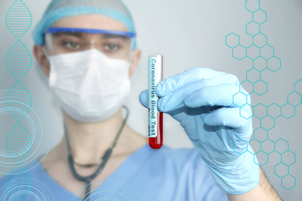 férfi orvos kék egyenruhában sztetoszkóppal védőmaszkban és szemüvegben néz egy laboratóriumi kémcső vérrel, koncepció a vizsgálat a vírus 2019-ncov - Fotó, kép