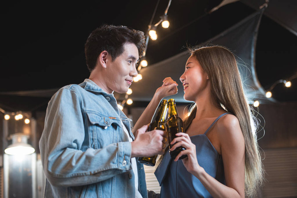 Genç Asyalı çift, gece partisinde dans edip içerek eğleniyor. Çatıdaki gece kulübünde ellerinde bira şişesi ve göz teması kurarak çiftler partisinde flört ediyor.. - Fotoğraf, Görsel