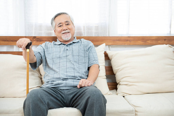 elderly old senior man sitting, resting his hands on wooden walking stick sitting on couch in living room in house after retirement. - Foto, imagen
