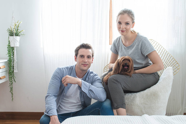 Young happy caucasian couple with smile sit on sofa  hug dog on knee in bedroom at home,indoors lifestyle concept. - Fotoğraf, Görsel