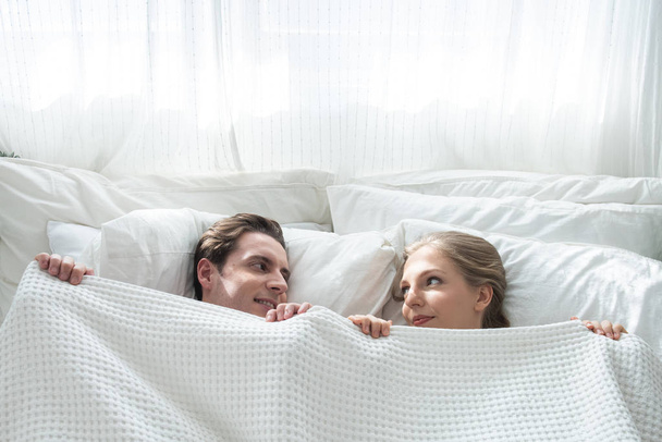 Κάτοψη της όμορφης νεαρό καυκάσιο ζευγάρι κρύβεται κάτω από κουβέρτα και κοιτάζοντας ο ένας τον άλλον με χαμόγελο και διασκέδαση, ενώ βρίσκεται στο κρεβάτι στο ξύπνημα πρωί στο σπίτι. - Φωτογραφία, εικόνα