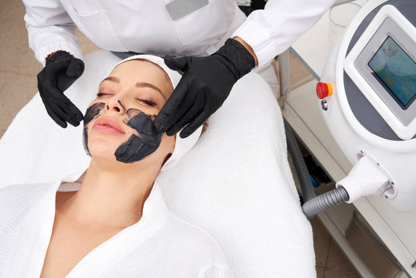 Cosmetologo applicare maschera nera sul viso di una bella donna per buccia di carbonio
 - Foto, immagini