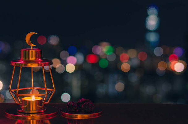 Moderní zlatá lucerna, která má na vrcholu měsíční symbol a malý talíř datlí ovoce s barevnými městskými bokeh světly pro muslimskou hostinu svatého měsíce ramadánu Kareem. - Fotografie, Obrázek
