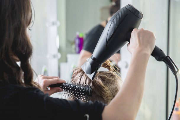 Крупный план рук парикмахеров, высушивающих длинные светлые волосы феном и круглой щеткой
 - Фото, изображение