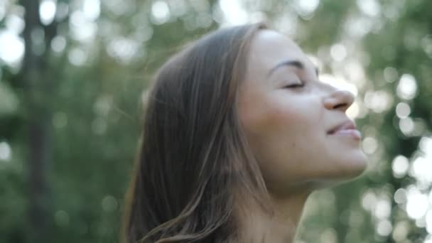 Close-up shot of an attractive smiling brunette girl with long hair, on which the breeze slightly blows. Focusing on the face of a young woman who enjoys the fresh air and nature of the park in spring - Metraje, vídeo