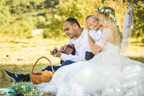 Picknick stijl buiten bruiloft. Bruidegom spelen op gitaar, jonge aantrekkelijke blomde bruid en kleine blonde jongen zittend op een deken in de zomer zonnige dag. - Foto, afbeelding